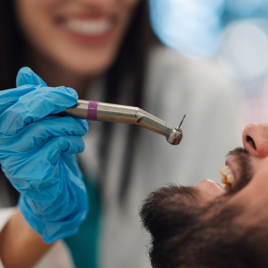 Smiling dentist wearing blue gloves holding a dental drill while performing a checkup on a male patient in a dental clinic, ensuring good oral hygiene